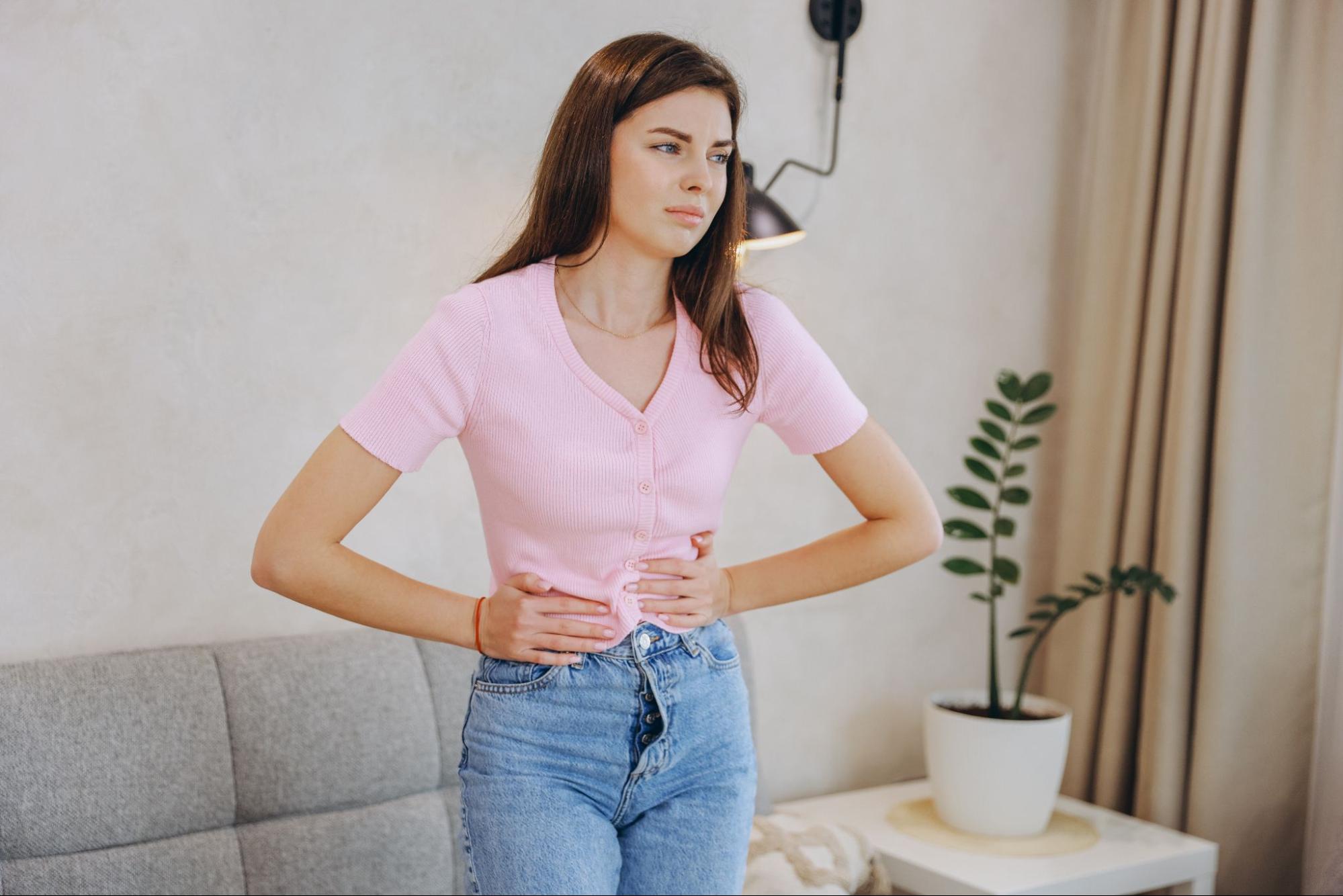Jovem segurando o estômago, lutando com dor abdominal enquanto está em sua sala, refletindo desconforto e angústia