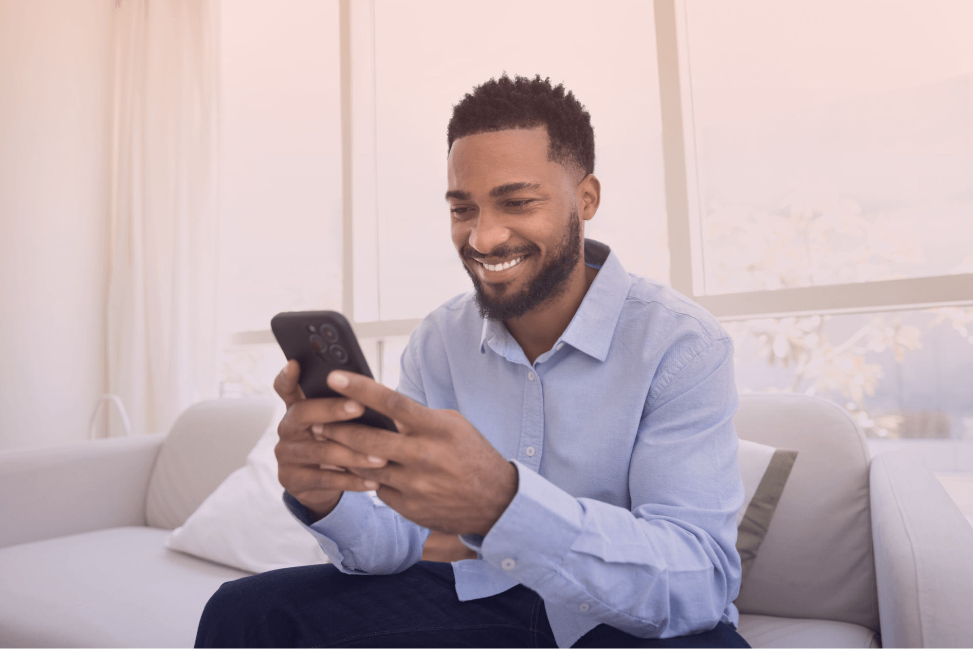 Homem jovem de pele escura e barba, vestindo uma camisa social azul clara, sorrindo enquanto usa um smartphone preto. Ele está sentado confortavelmente em um sofá branco em um ambiente moderno e bem iluminado, com grandes janelas ao fundo, transmitindo uma sensação de relaxamento e conectividade digital.