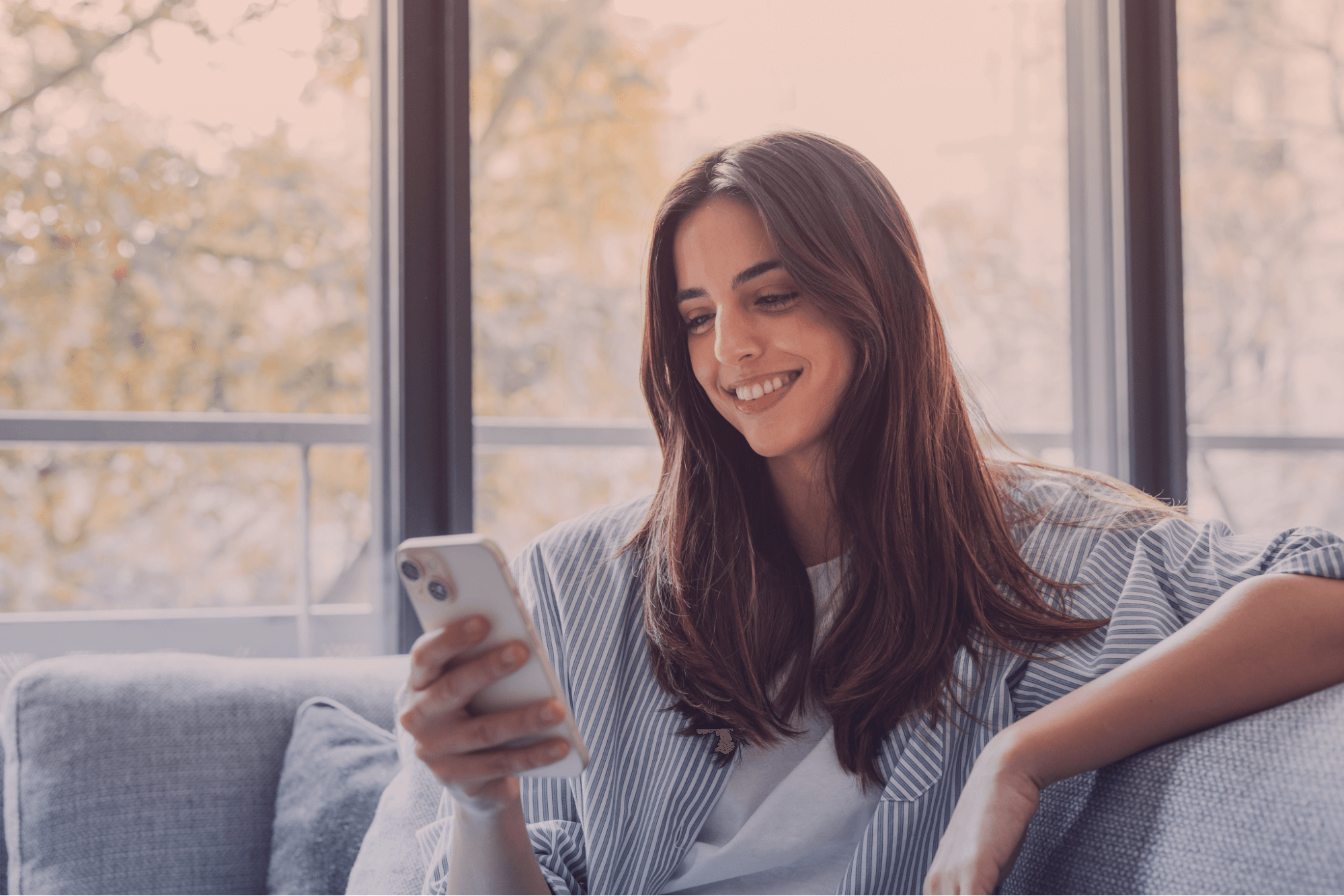 Mulher jovem sorridente com cabelos castanhos longos, usando um smartphone branco enquanto relaxa em um sofá cinza. Ela está em um ambiente interno bem iluminado, com uma janela grande ao fundo, transmitindo uma sensação de conforto e conexão digital.