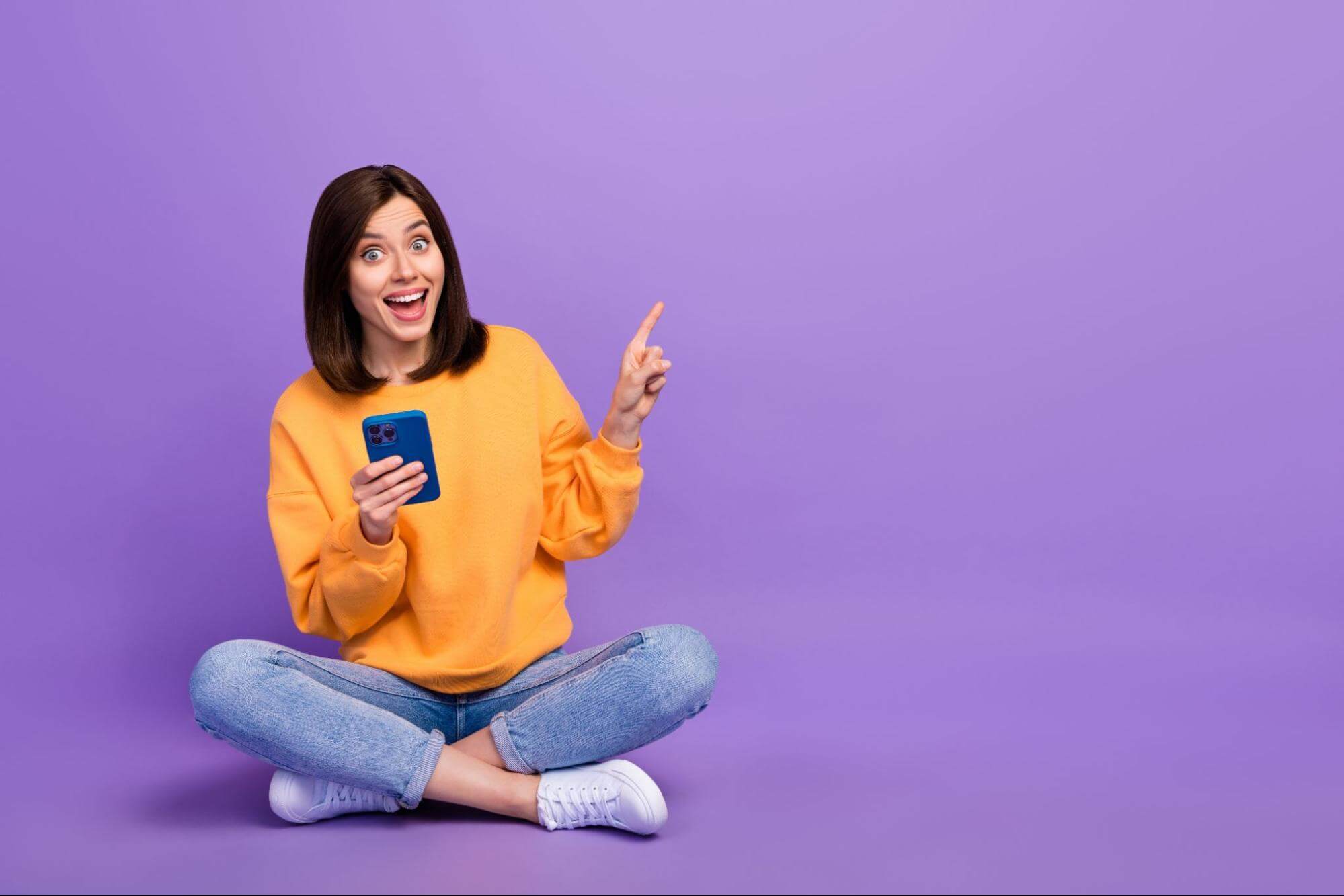  Mulher jovem e animada com cabelo castanho, vestindo um moletom laranja e jeans, sentada de pernas cruzadas no chão. Ela segura um smartphone azul e aponta com o dedo indicador para o lado direito, com uma expressão de surpresa e alegria em um fundo roxo vibrante. Ideal para campanhas de marketing, promoções de aplicativos ou anúncios de produtos digitais.