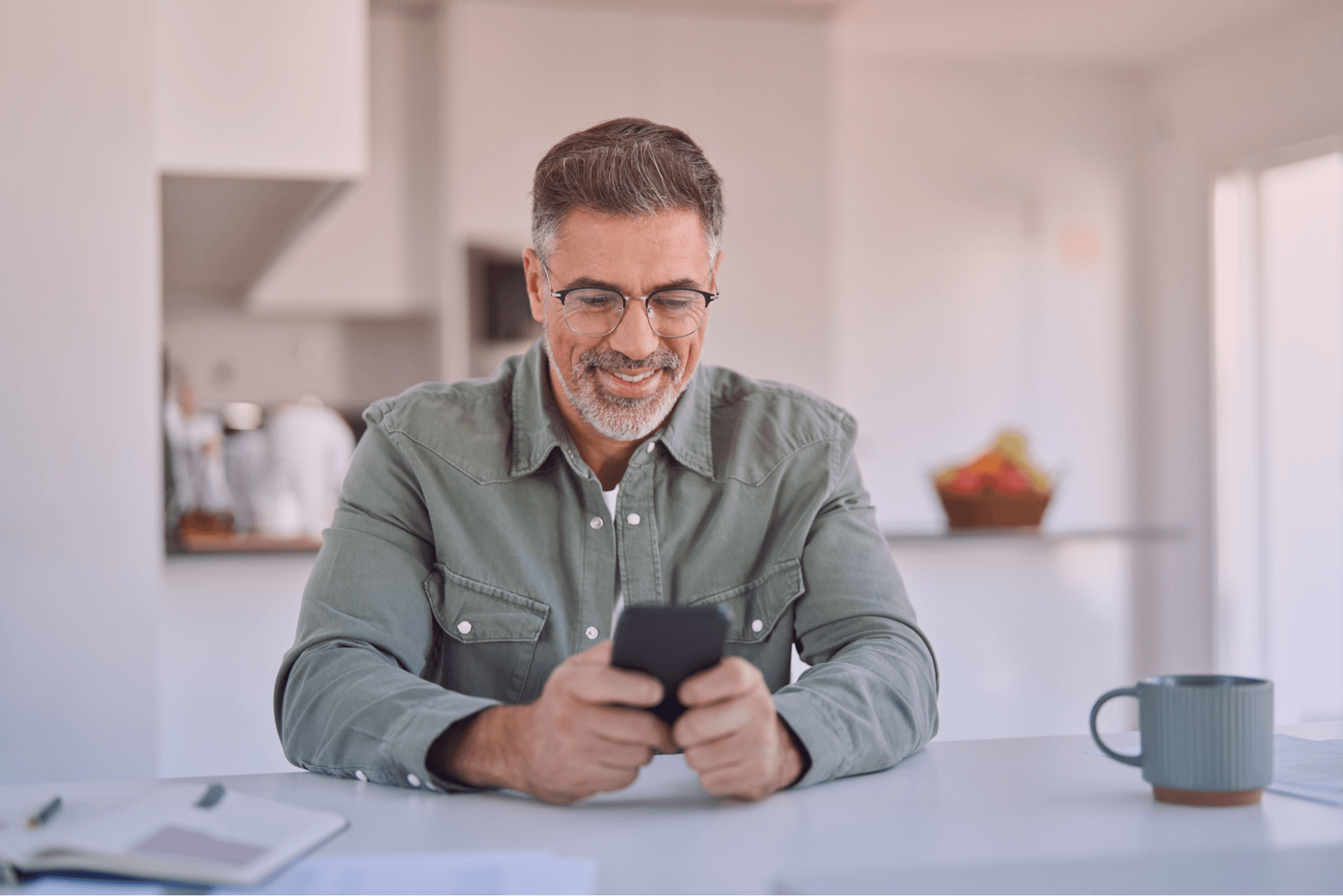 Homem de meia-idade sorrindo, com cabelo grisalho e barba, usando óculos e uma camisa verde-oliva, enquanto interage com um smartphone preto. Ele está sentado em uma mesa branca com uma caneca e documentos, em um ambiente doméstico moderno e iluminado, transmitindo uma sensação de relaxamento e conectividade.