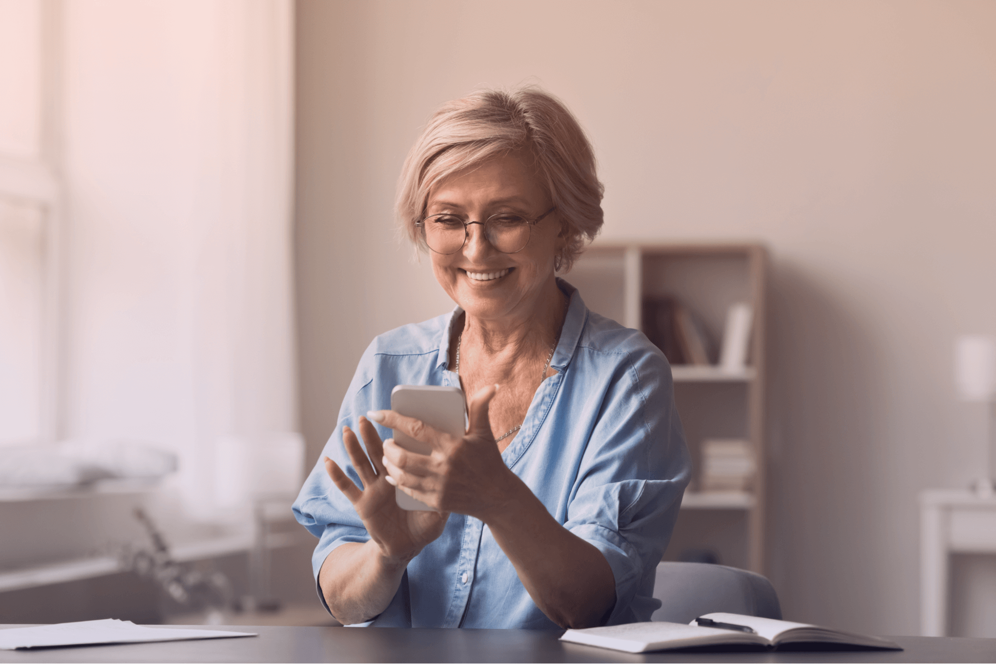 Mulher idosa feliz com cabelo loiro e óculos, sorrindo e interagindo com um smartphone branco. Ela está sentada em uma mesa de escritório, com um caderno e caneta, em um ambiente de trabalho iluminado, transmitindo satisfação ao usar tecnologia.