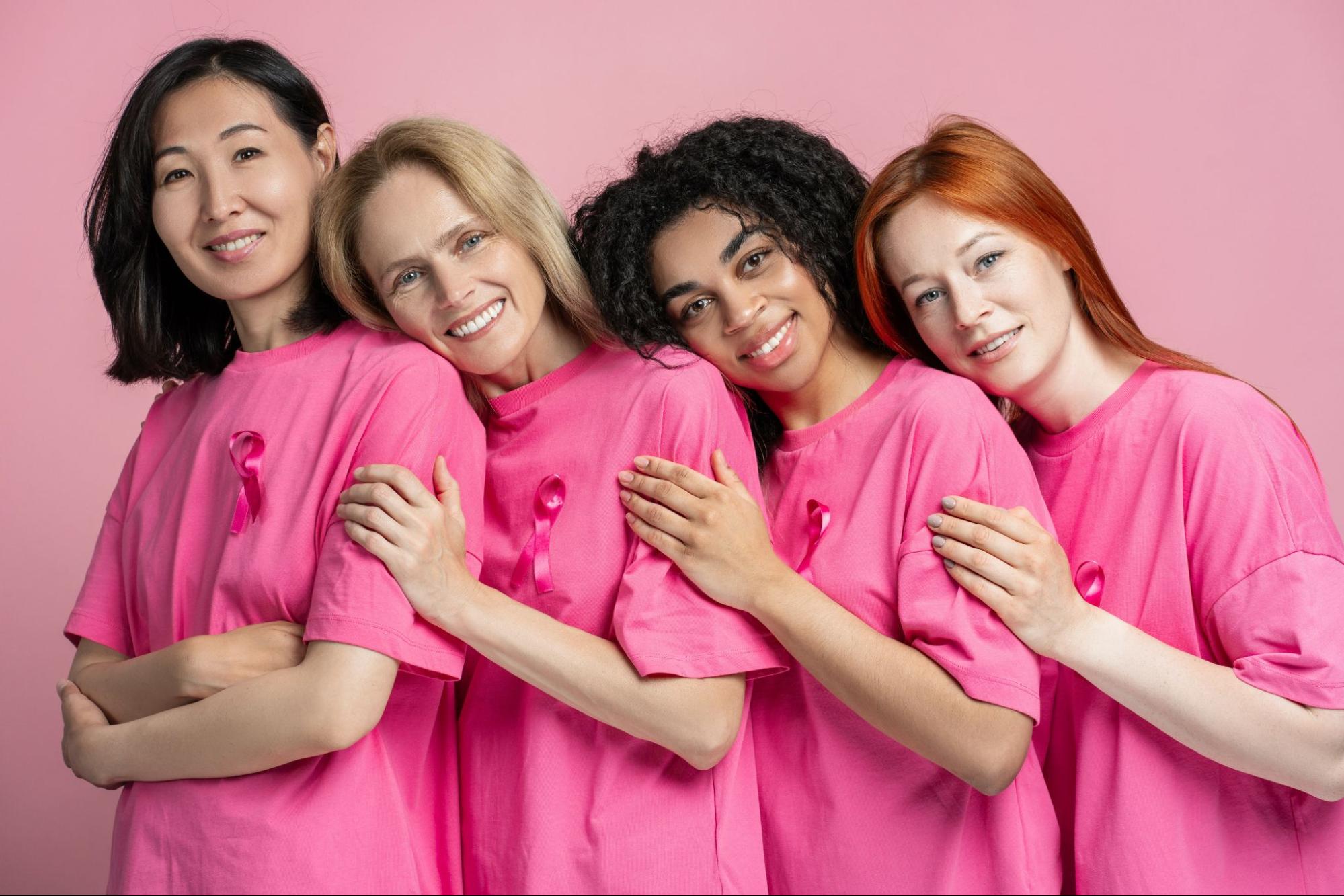 Um grupo diversificado de mulheres de diferentes idades e etnias estão juntas, vestindo camisetas rosa com fitas rosa, simbolizando seu apoio à conscientização sobre o câncer de mama.
