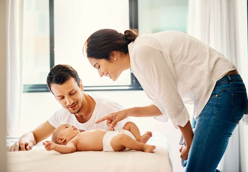 Casal olha e brinca com bebê deitado em uma cama