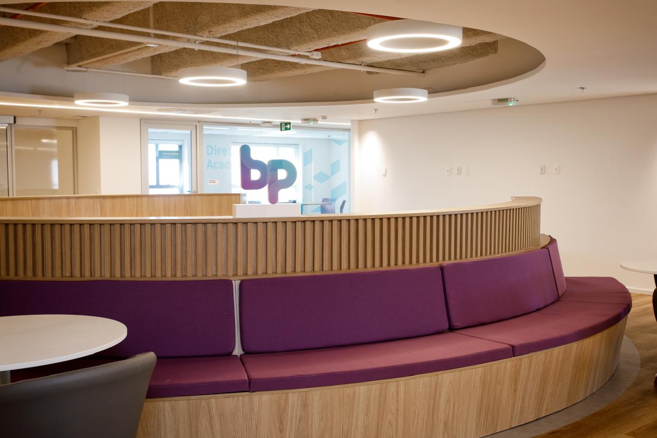Ambiente moderno na Faculdade BP, com um banco circular de madeira clara e almofadas roxas, paredes brancas e o logo da faculdade ao fundo
