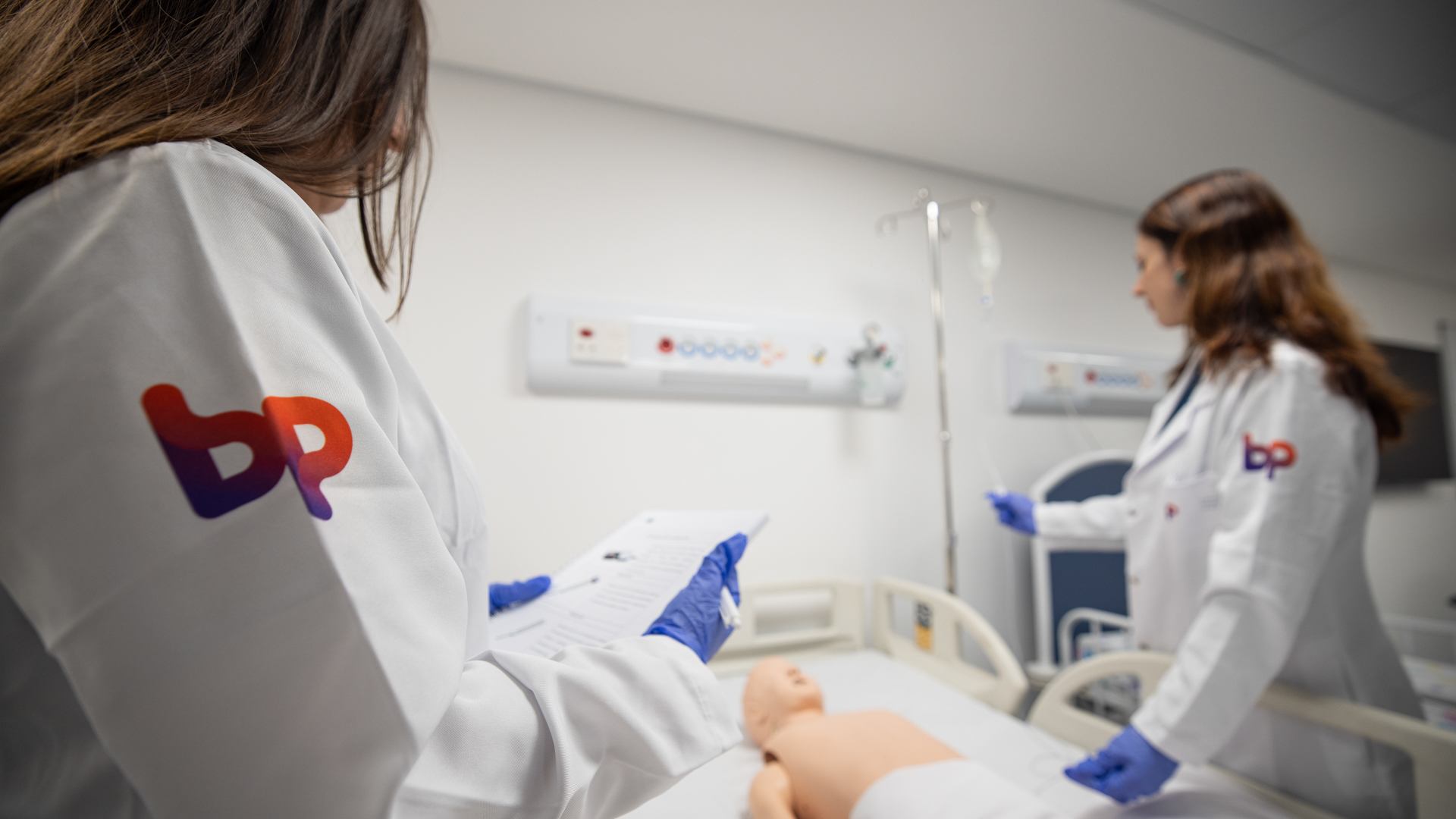 Duas profissionais com jalecos da BP realizam uma atividade prática em ambiente de simulação hospitalar, com manequim de treinamento em um leito.