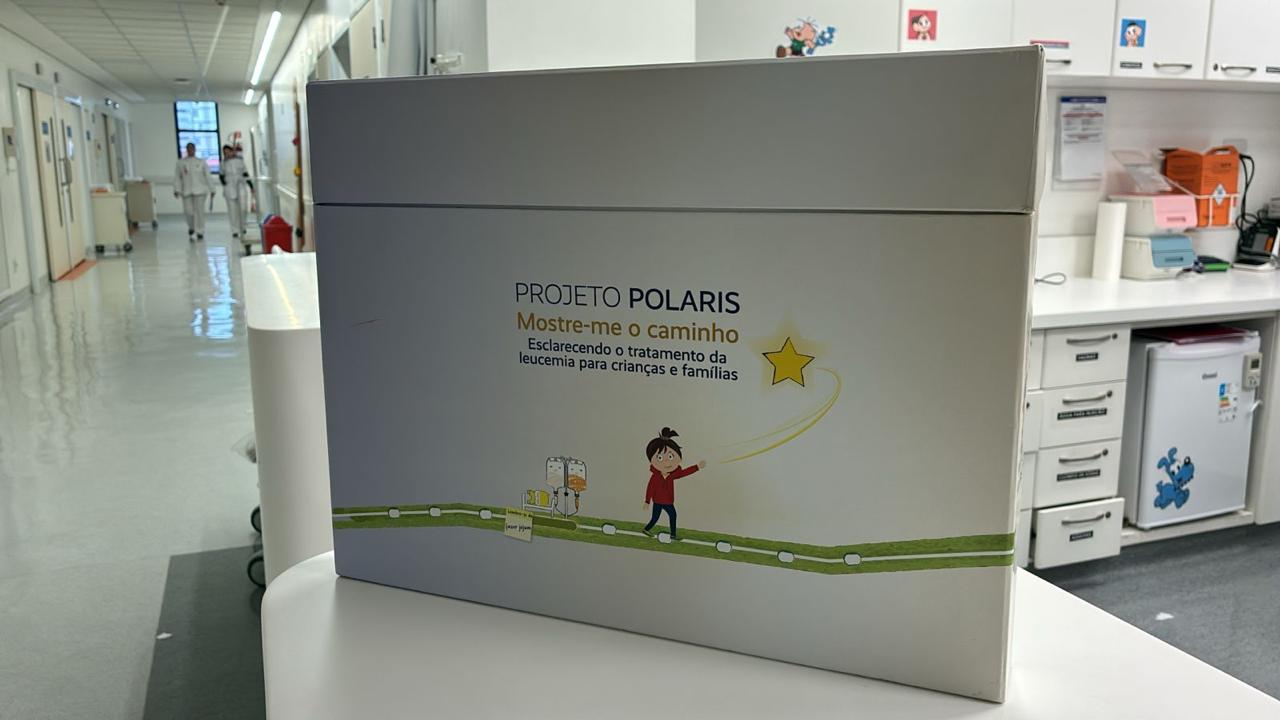 Imagem de um ambiente interno do Hospital BP, com uma grande placa mostrando um desenho lúdico de uma criança explicando o tratamento de leucemia. Na placa, está escrito "Projeto Polaris - Mostre-me o caminho: Esclarecendo o tratamento da leucemia para crianças e famílias