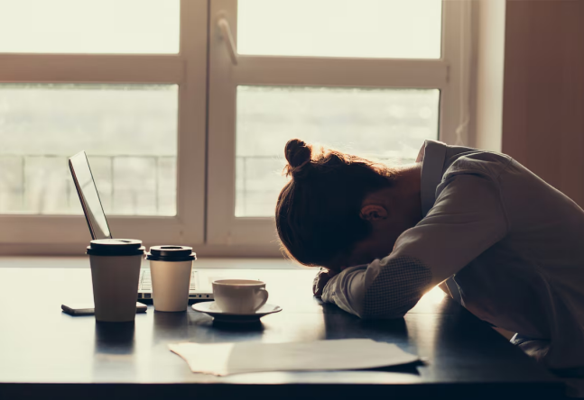 Uma mulher de cabelo preso em um coque encostada com os braços em cima de uma mesa e com a cabeça baixa, como se estivesse cansada. Na sua frente tem vários copos de café e um notebook.