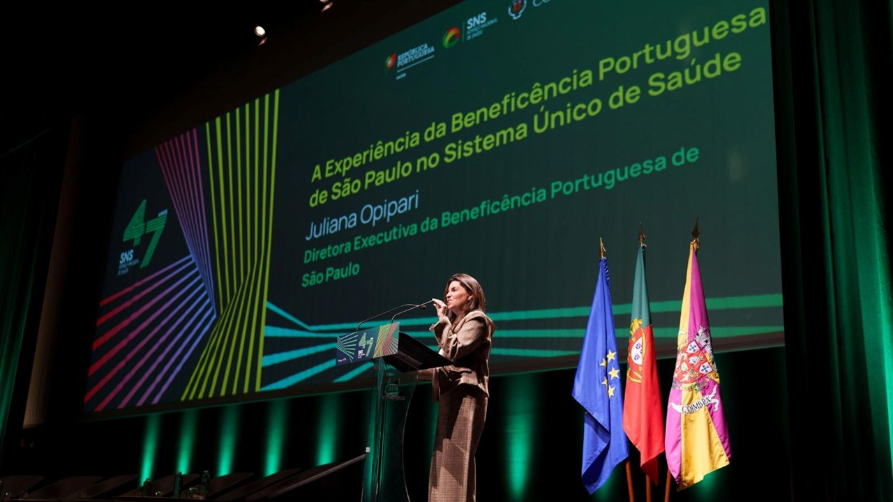 Mulher fala ao microfone em um púlpito, sobre um palco, diante de um grande telão. Na tela, aparece o título “A Experiência da Beneficência Portuguesa de São Paulo no Sistema Único de Saúde” e o nome Juliana Opipari. Ao lado do púlpito estão as bandeiras da União Europeia, de Portugal e de Coimbra.
