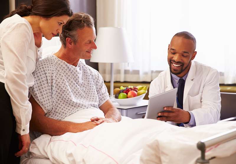 Médico insere informações em um tablet enquanto conversa com paciente internado e sua acompanhante.